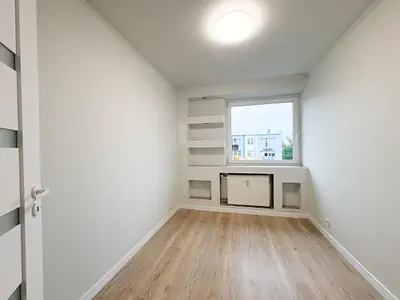 Mały pusty pokój z jasną podłogą, wbudowanymi półkami przy oknie i kaloryferem.