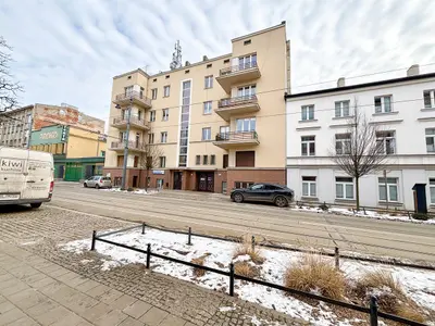 Widok ulicy na beżowy apartamentowiec z balkonami, torami tramwajowymi i samochodami przed budynkiem