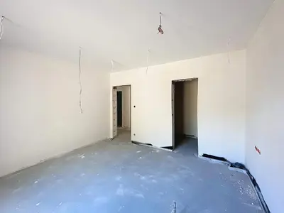 Duże pomieszczenie w stanie surowym, betonowa podłoga i dwa przejścia do kolejnych pokoi.