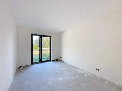 Kolejny pokój w stanie surowym z betonową podłogą i drzwiami balkonowymi na zieleń.