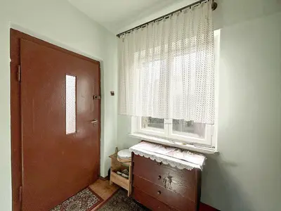 Małe wejście z brązowymi drzwiami, oknem z koronkowymi zasłonami i drewnianą komodą pod oknem.