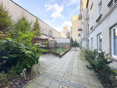 Wybrukowane podwórko z zielenią wzdłuż jasnego budynku kamienicy na Bałutach